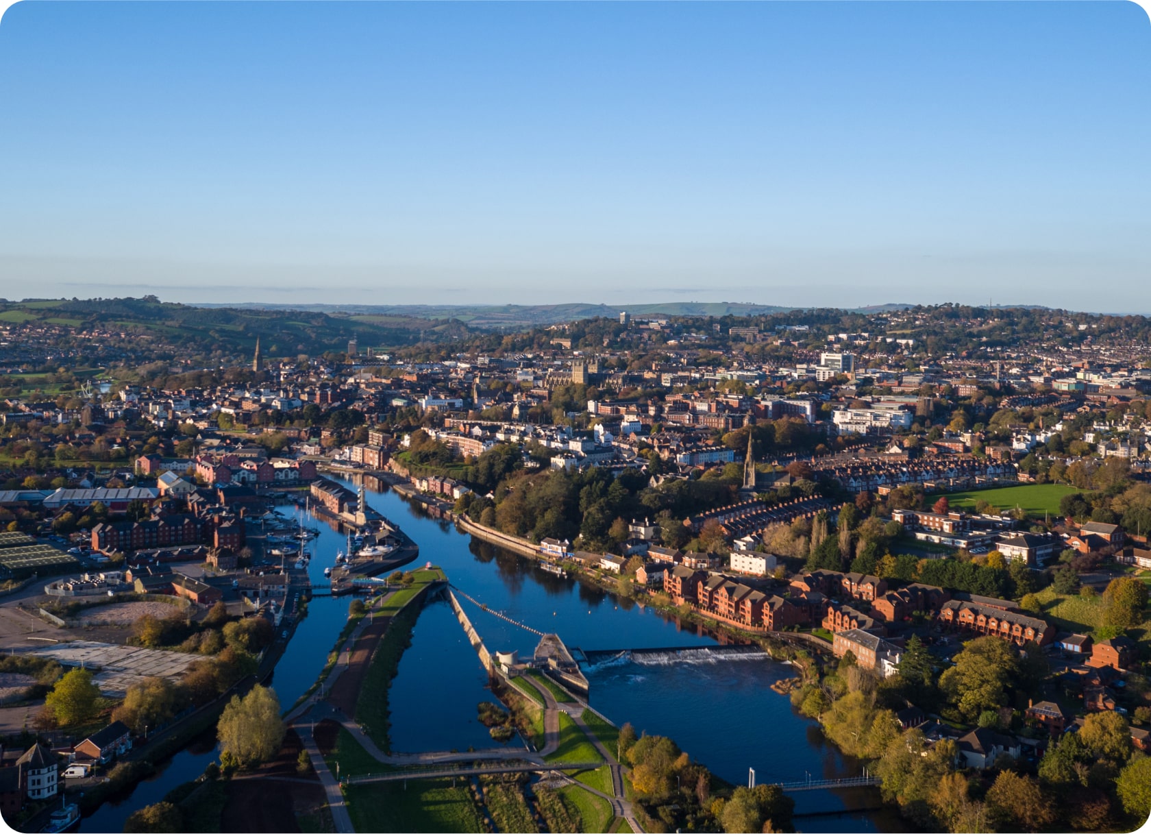 Aerial view of a city with winding river cutting through the landscape. The city features numerous buildings, bridges, and lush green areas. The horizon shows distant hills under a clear blue sky, and the scene is bathed in sunlight, highlighting the vibrant colors.