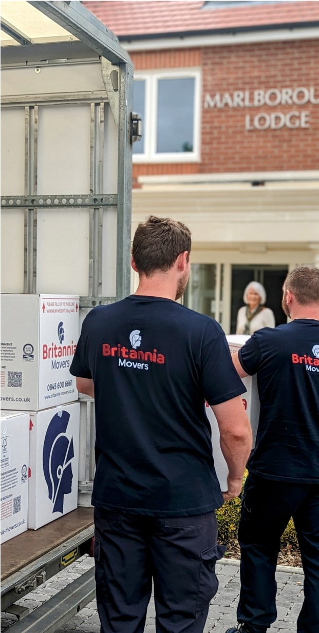 Two movers in black Britannia Movers shirts carry boxes labeled "Britannia Movers" from a truck towards the entrance of Marlborough Lodge. A woman stands at the entrance observing them.