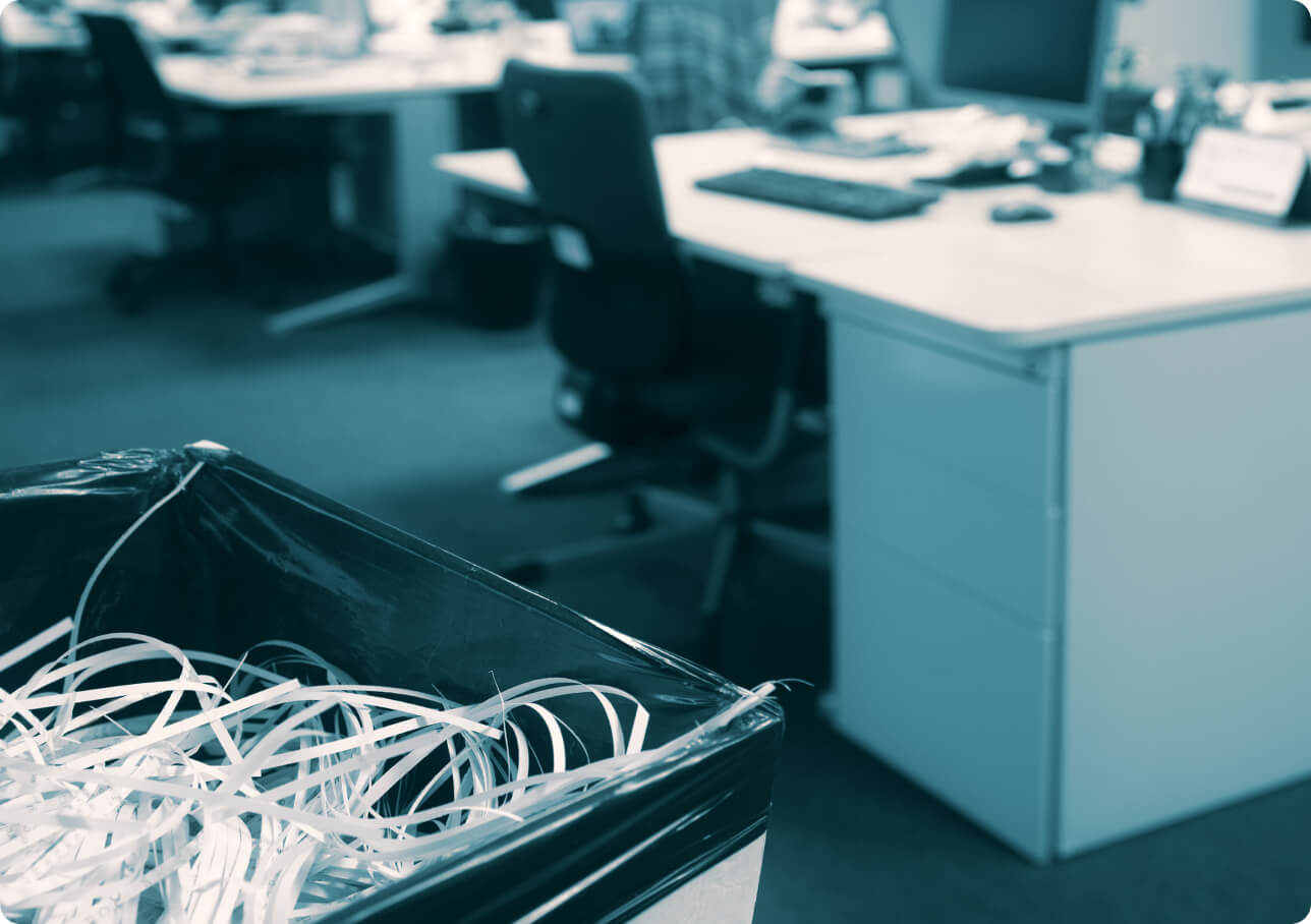 A close-up of a trash bin filled with shredded paper in an office setting. In the background, there are desks with computer monitors, chairs, and office supplies, slightly out of focus, giving a sense of a busy work environment.