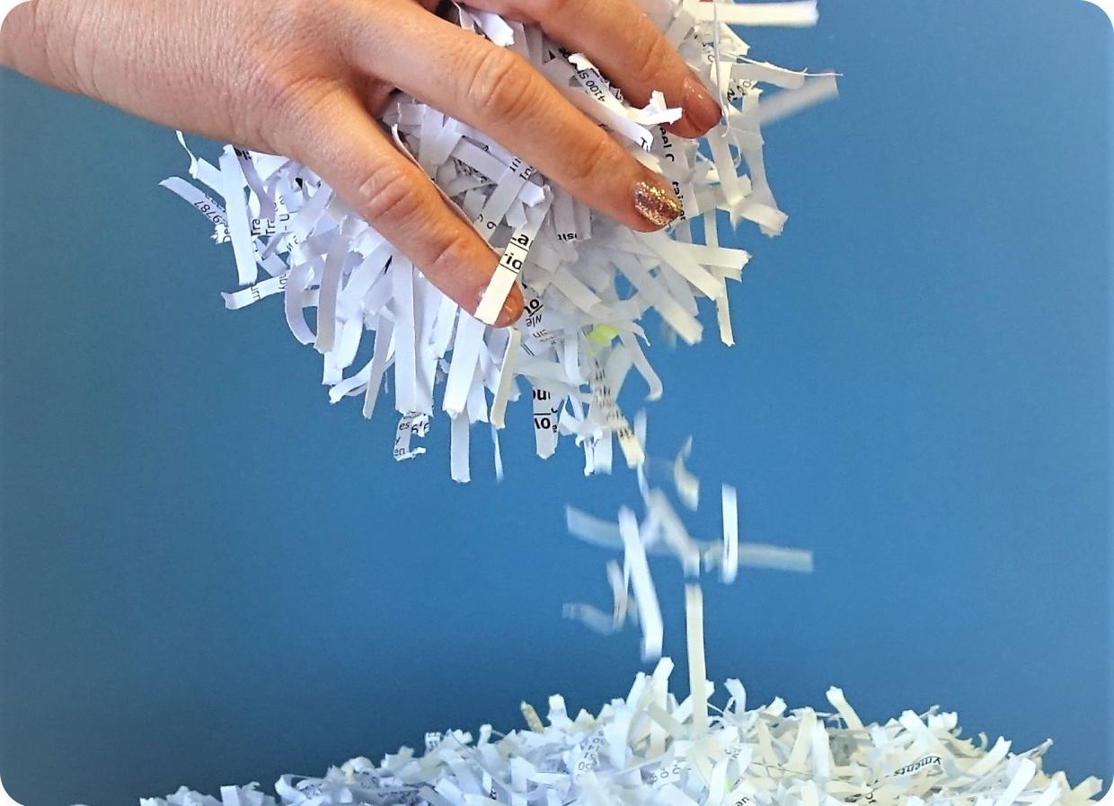 A hand releasing shredded paper over a pile of shredded paper, against a blue background. The shredded strips are white with some visible text fragments. The fingernails of the hand are painted with glittery nail polish.