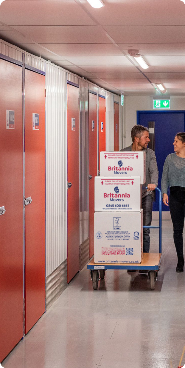 A man and woman are walking in a hallway with red doors, pushing a blue cart loaded with several white boxes labeled "Britannia Movers." The hallway appears to be part of a storage or moving facility, with the boxes secured on the cart.