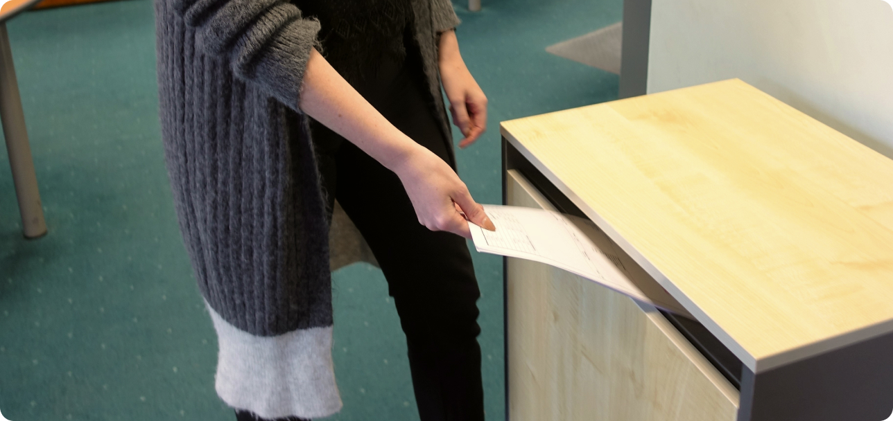 A person wearing a grey sweater and black pants is inserting a paper into a light wood and grey metal collection box. The person is standing in a room with a blue carpet.