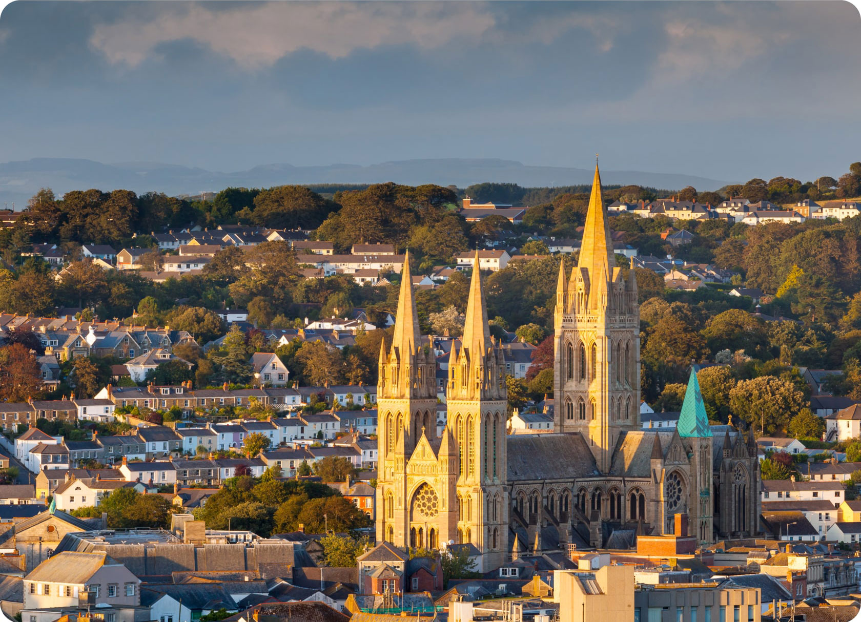 A grand cathedral with tall spires and intricate architecture stands prominently in the foreground, surrounded by a town with numerous houses and lush green trees. The lighting suggests it's either early morning or late afternoon, casting a warm glow on the scene.