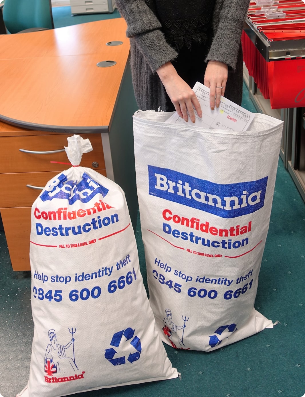 A person is placing documents into two large white sacks labeled "Confidential Destruction Britannia." The bags feature blue and red text and include a recycling symbol and a phone number for help with identity theft. The scene appears to be an office setting.