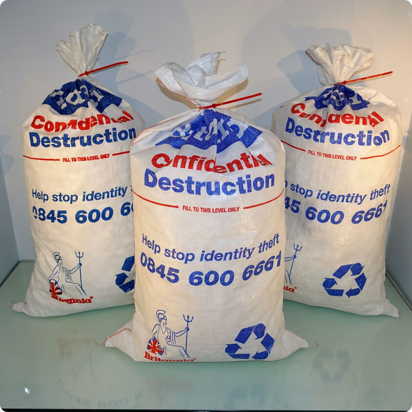 Three white sacks labeled "Confidential Destruction" with red and blue text are displayed. They have recycling symbols and a phone number, 0845 600 6661, printed on them. The bags are tied at the top and placed on a glass surface.