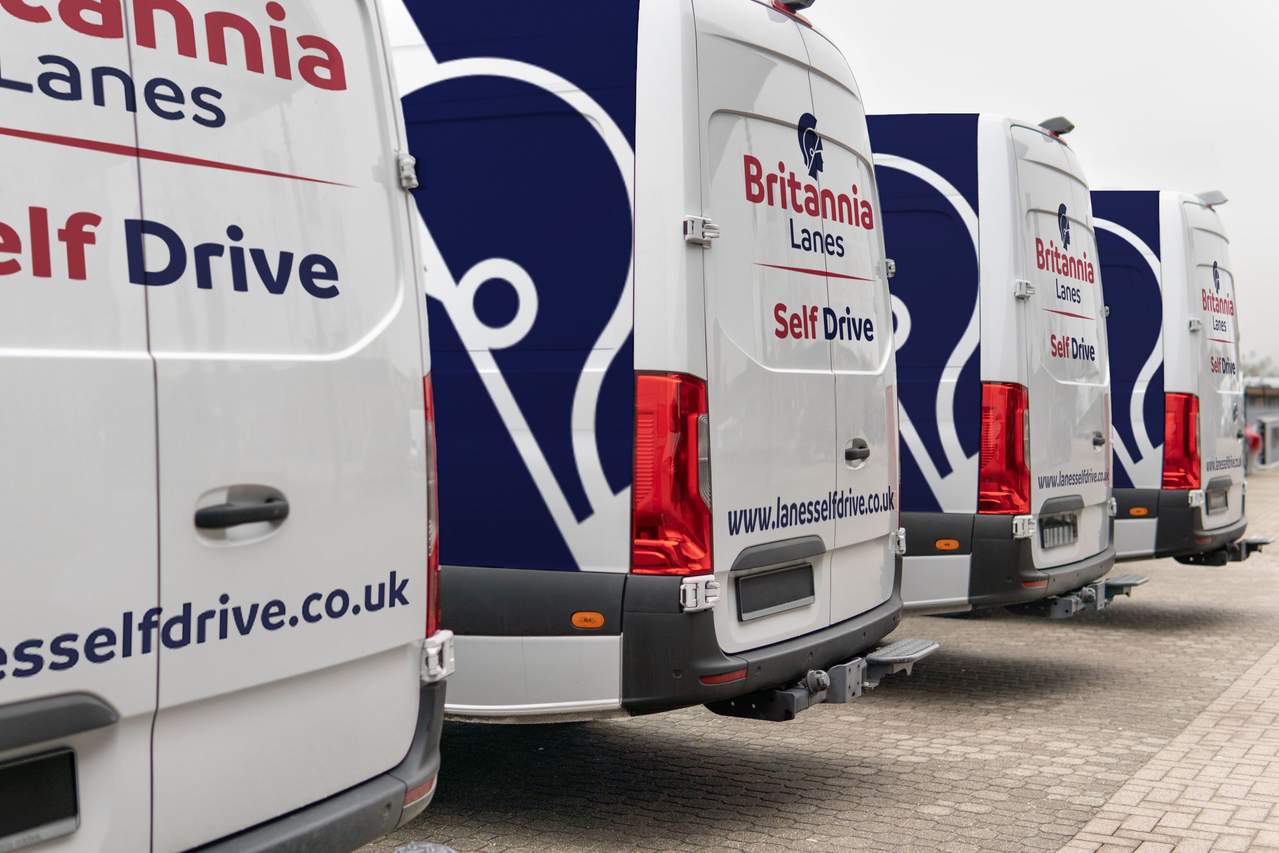 A row of Britannia Lanes Self Drive vans parked in a lot. The vehicles are white with dark blue accents and the company name, logo, and website (lanesselfdrive.co.uk) printed on the sides and back doors. The ground is paved with light-colored bricks.