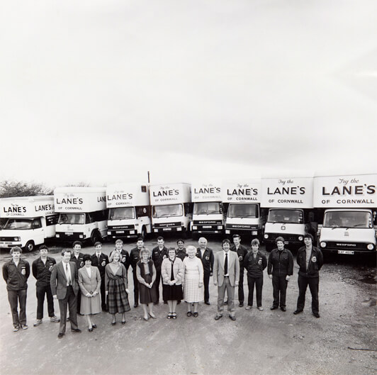 A black and white photo shows a line of delivery trucks labeled "Lane's of Cornwall" with their drivers and employees standing in front. The group is composed of men and women of varying ages, all posing and smiling at the camera in an open lot.