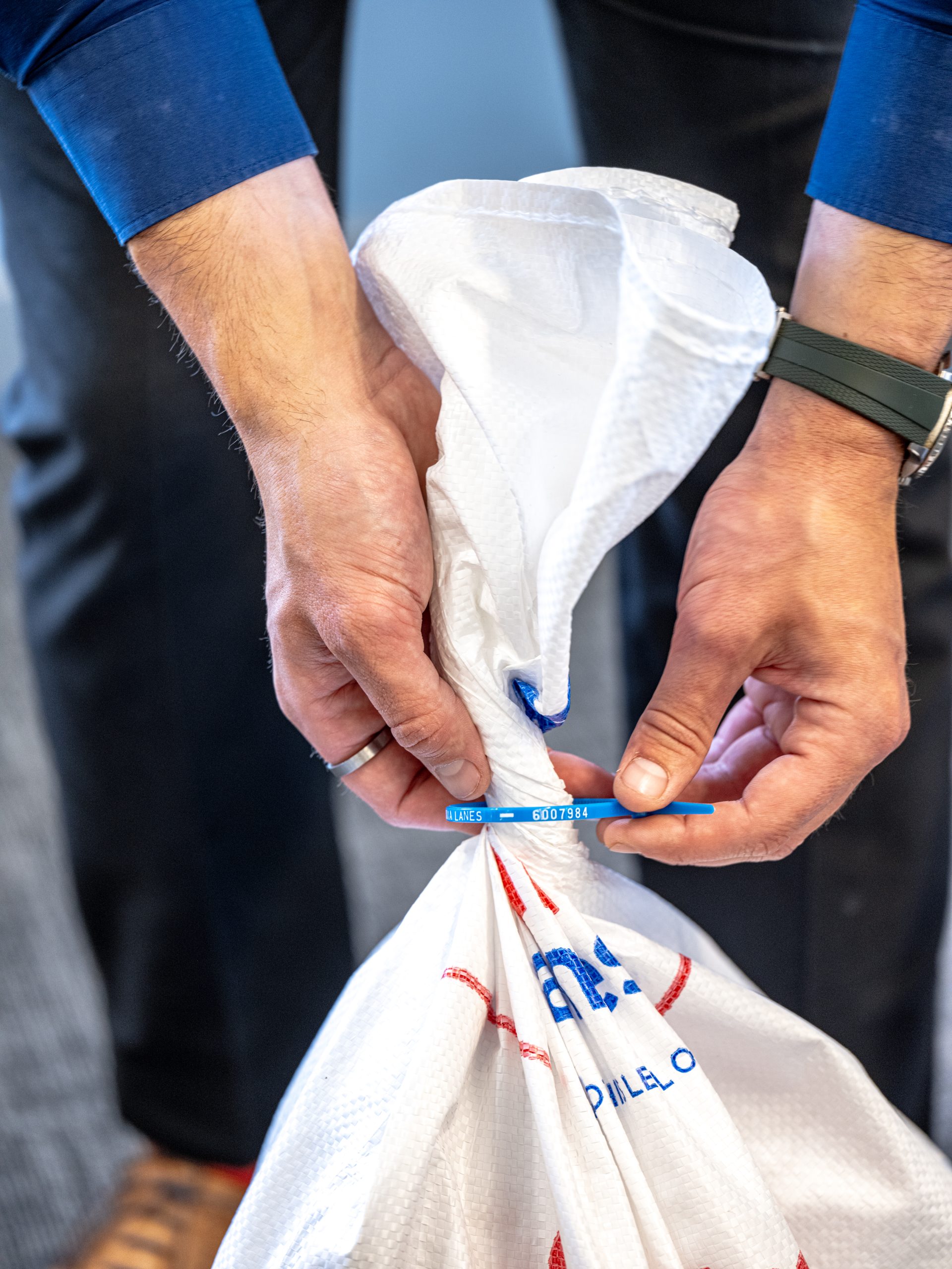 A person wearing a blue long-sleeve shirt and a wristwatch is securing a white sack with a blue zip tie. The sack has some red and blue markings on it. The person's hands are in the center, focusing on tightening the zip tie.