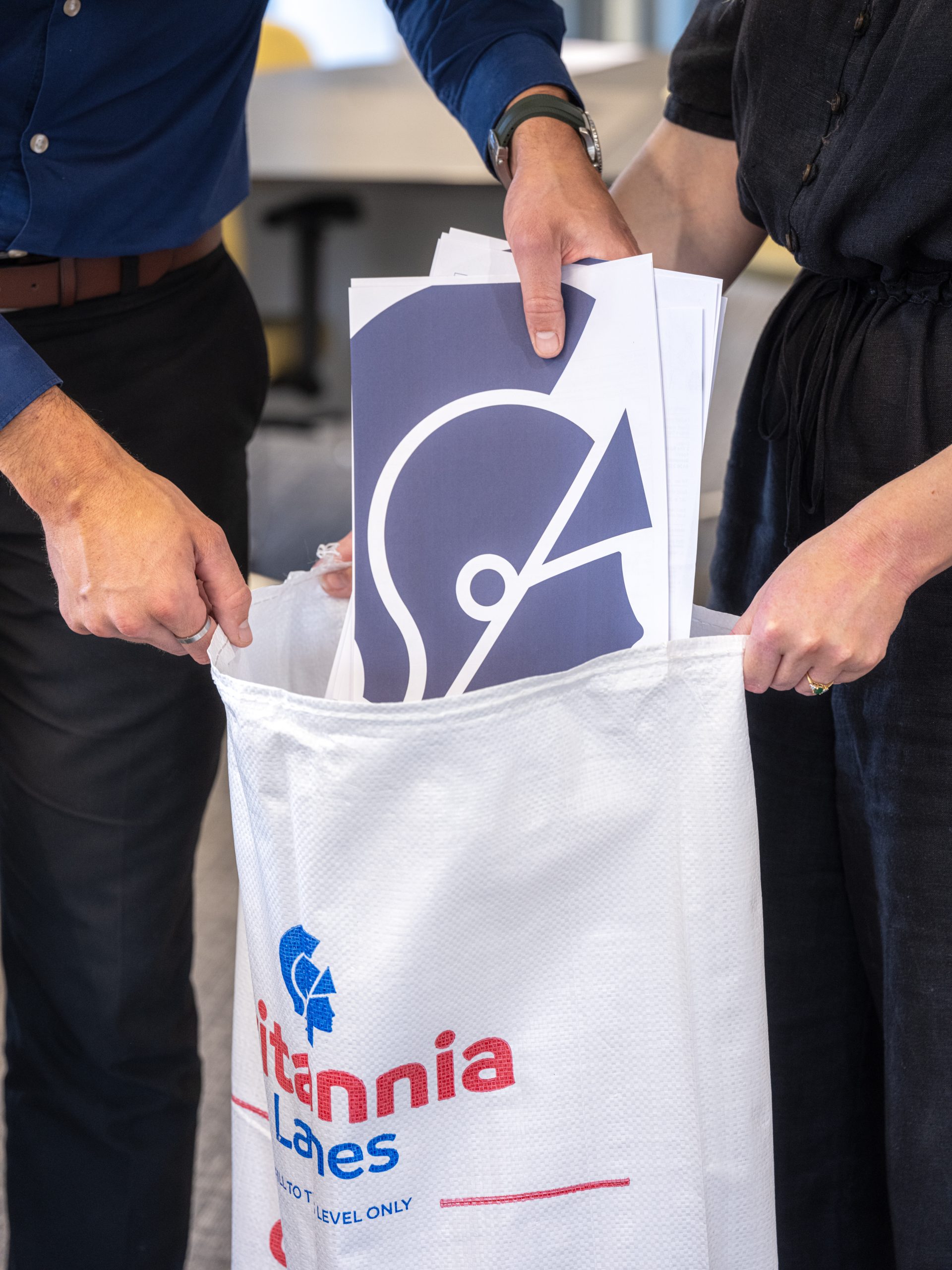 Two people are seen inserting documents into a large collection bag branded with "Britannia Lanes". One of the documents features a blue icon resembling an abstract face in profile. The scene appears to be in an office setting.