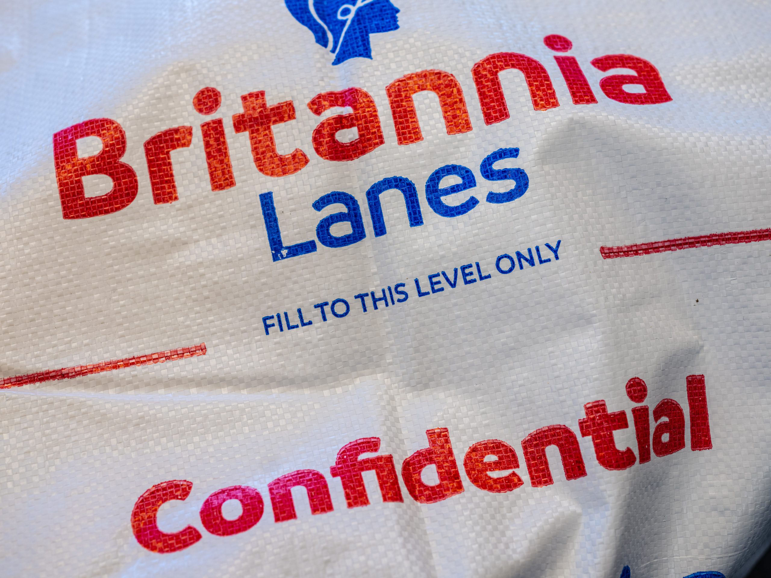 A close-up view of a white sack with "Britannia Lanes" written in red and blue letters. Below it, instructions say "Fill to this level only." The word "Confidential" is printed in large red letters near the bottom of the sack.