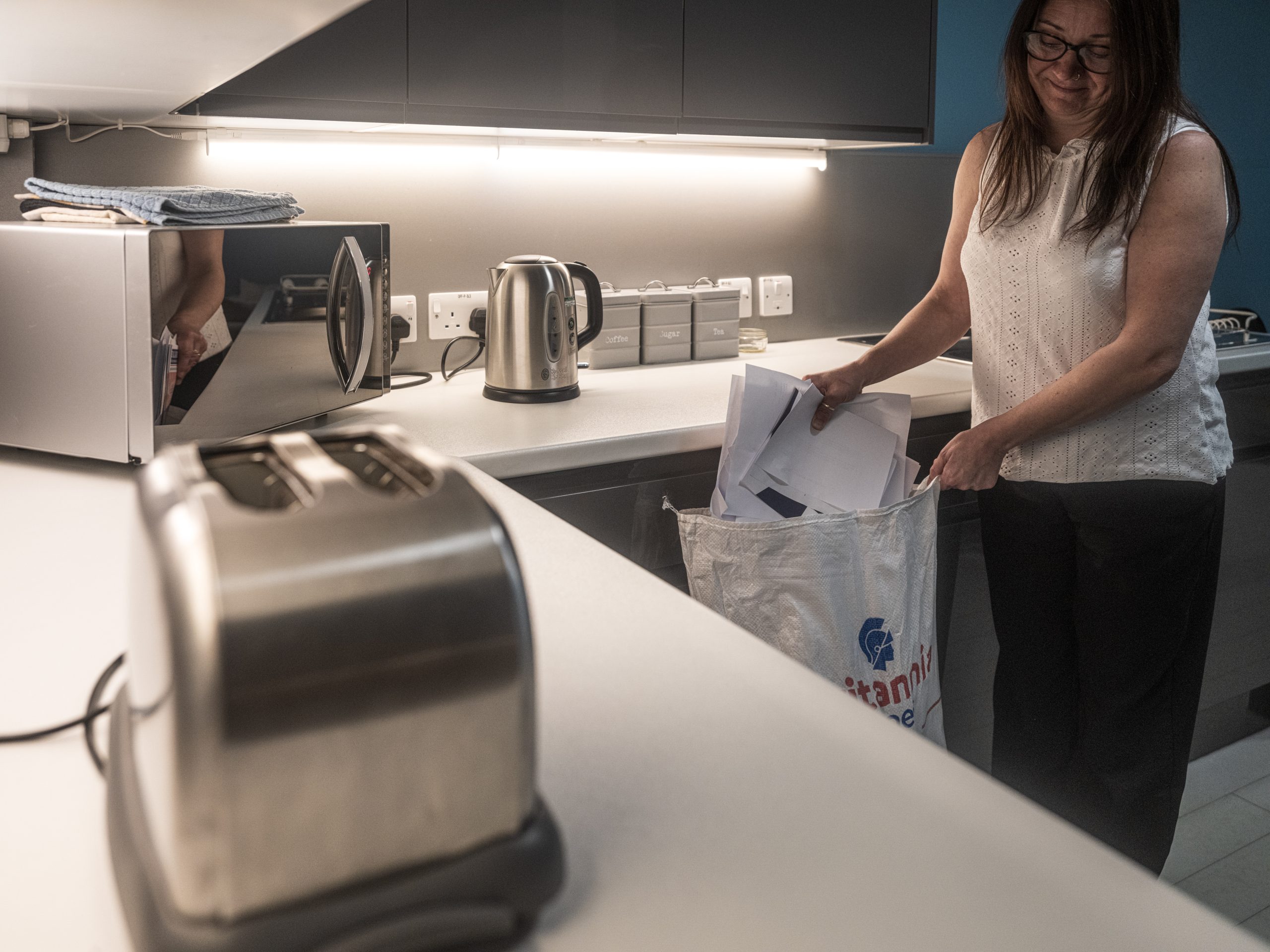 A woman in a kitchen is holding a bag while placing items into it. The countertop features a toaster, a kettle, and a microwave. Under-cabinet lighting illuminates the countertop area. The kitchen has a clean and modern appearance.