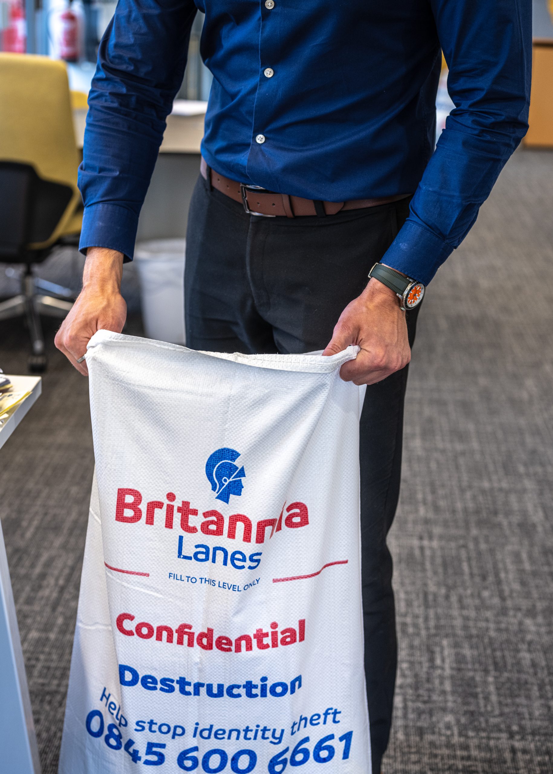 A person wearing a blue shirt and a watch is holding open a white sack labeled "Britannia Lanes" for confidential document destruction. The sack includes contact details and a reminder to help stop identity theft. The setting appears to be an office.