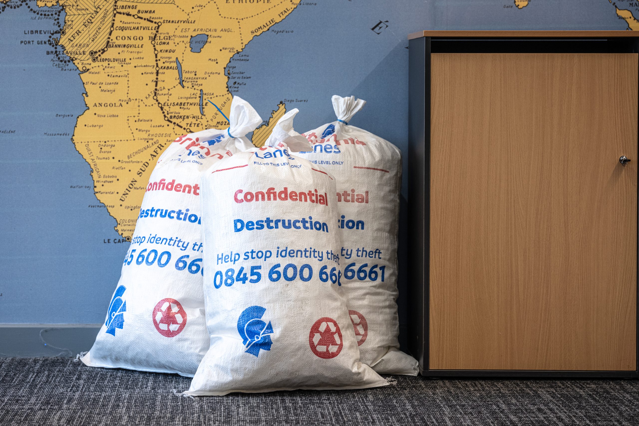 Three large white bags labeled "Confidential Destruction" and displaying a recycling symbol are placed near a wooden cabinet. They are positioned in front of a wall with a world map. Text on the bags includes a helpline number for preventing identity theft.