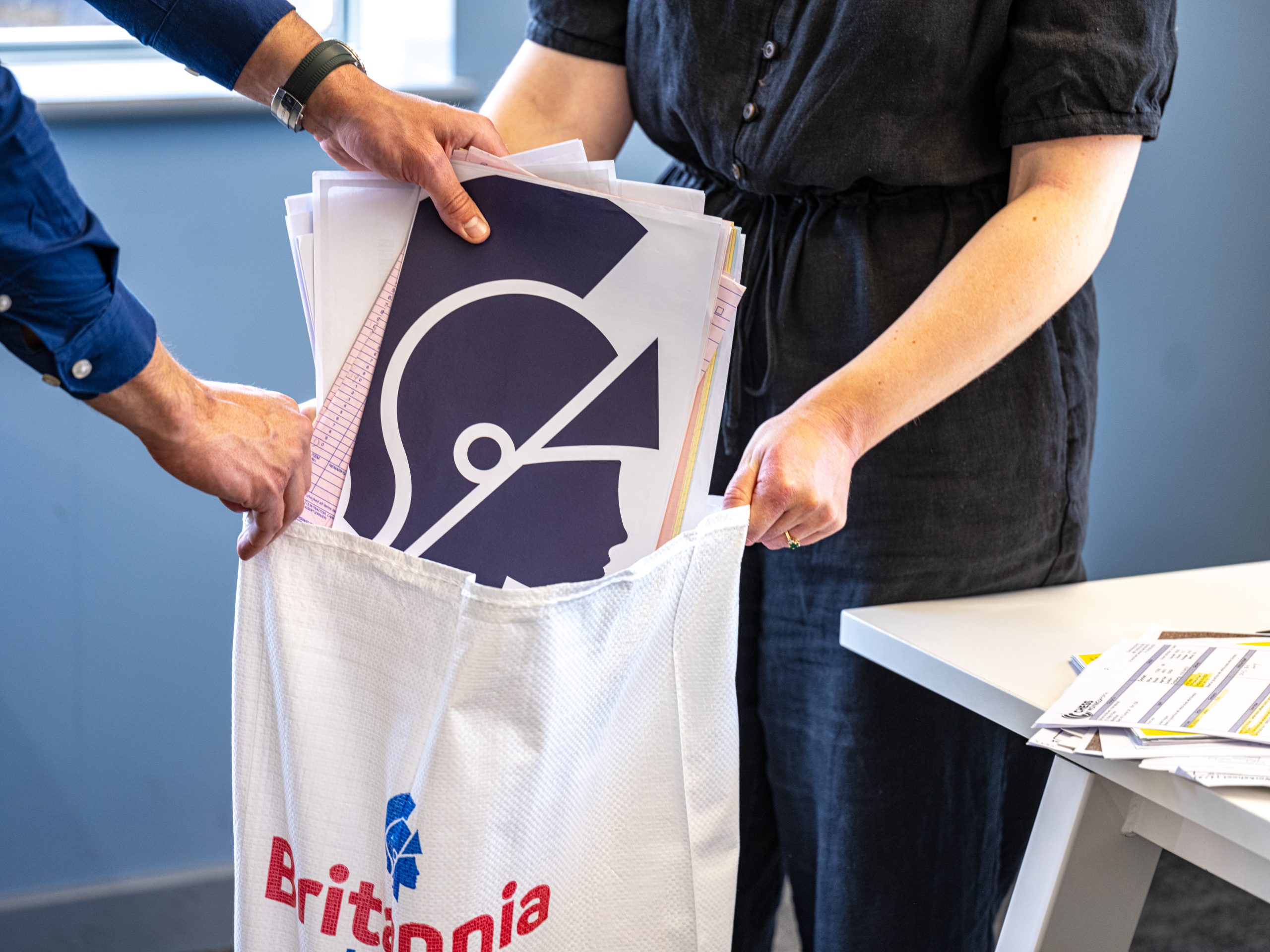 Two people are placing papers into a white bag that has "Britannia" printed on it in red and blue letters. The papers include a large illustration of a helmeted figure. The individuals' faces are not shown. A table with more papers is partially visible on the right.