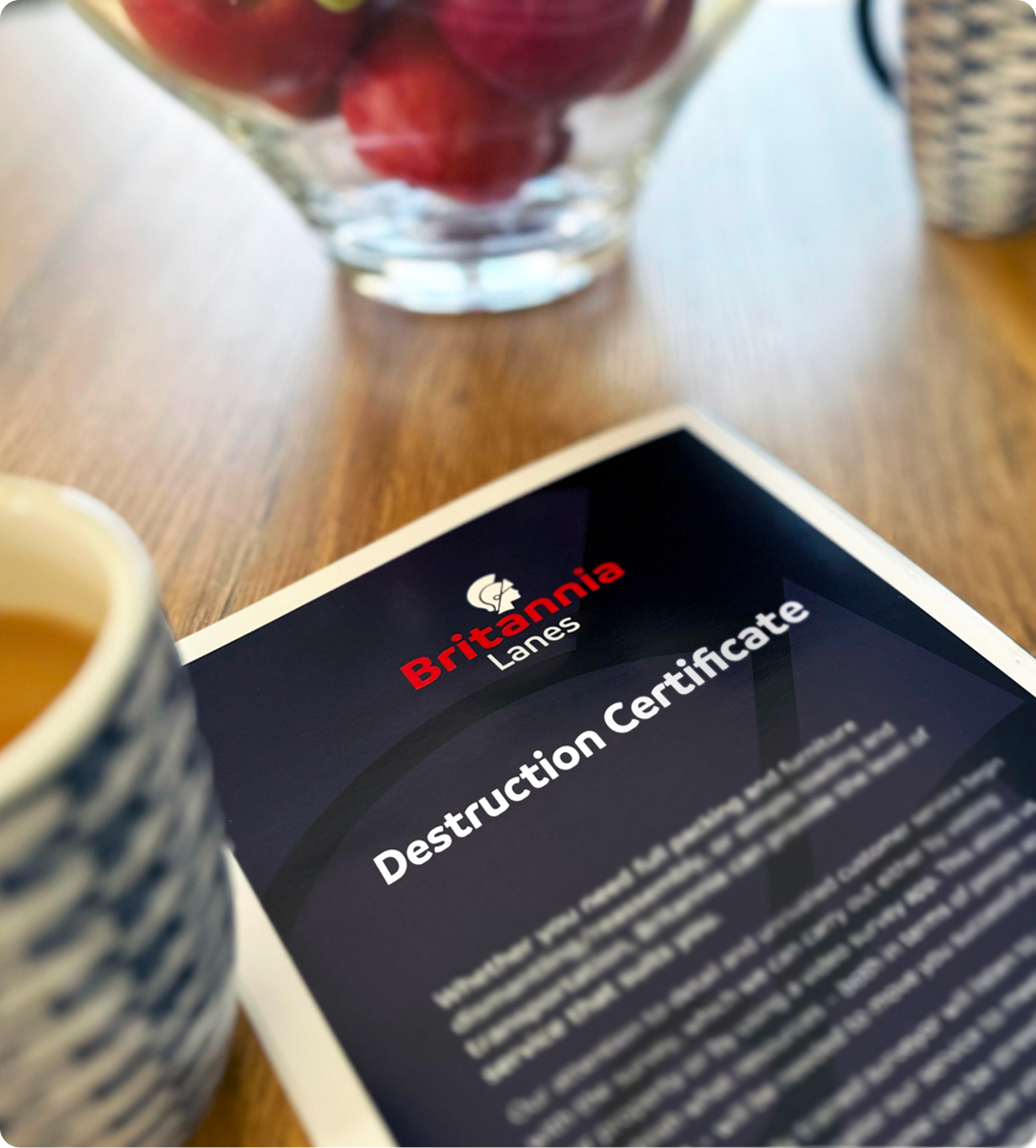 A document titled "Destruction Certificate" with the header "Britannia Lanes" lies on a wooden table. Nearby, there is a ceramic cup with a blue and white pattern and a glass bowl filled with red apples. The background is slightly blurred.