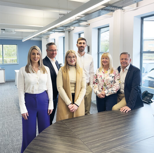 A group of six people, four men and two women, stand and sit around a conference table in a modern office space. They are smiling and wearing business-casual attire. The room has large windows and ample natural light.