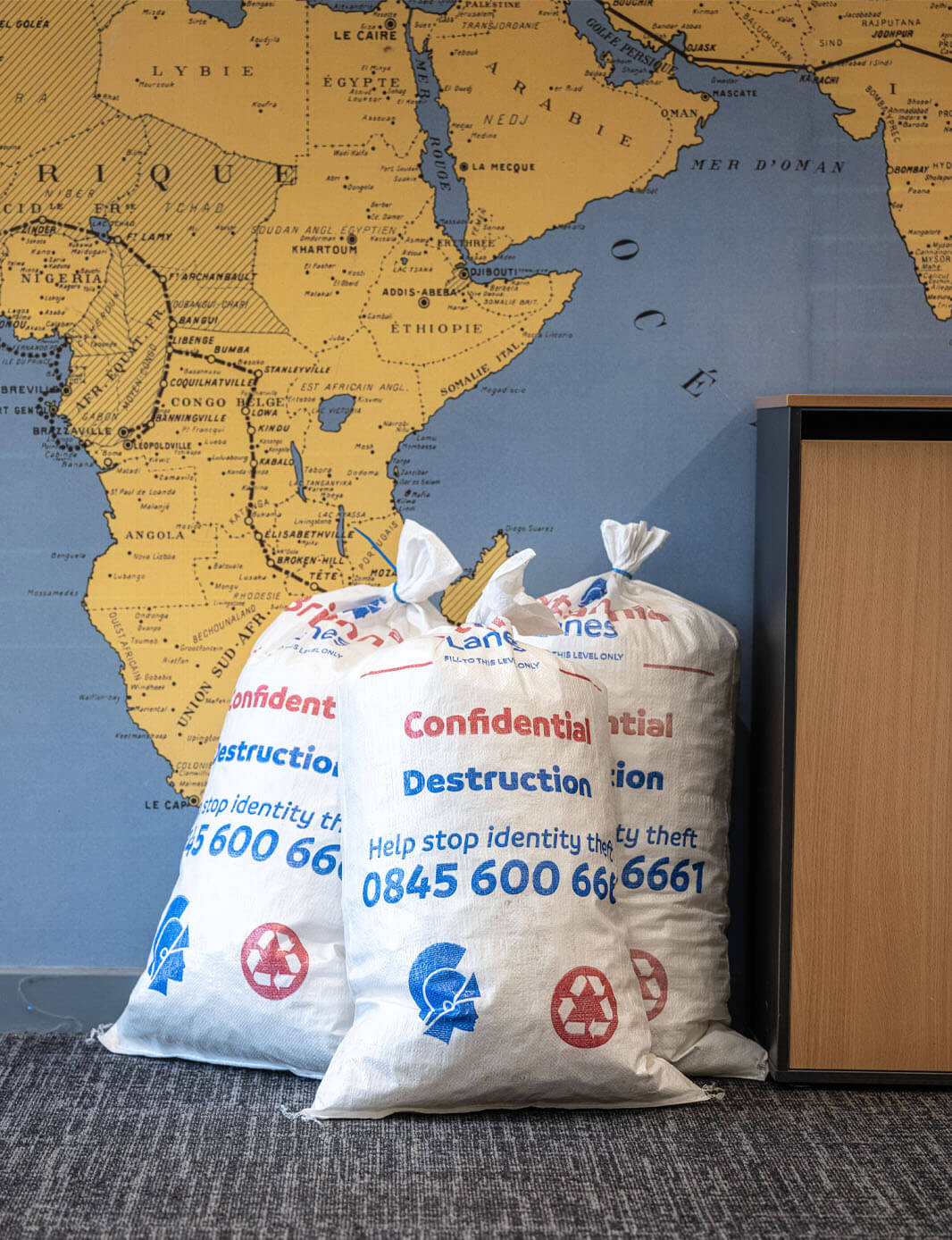 Three white sacks labeled “Confidential Destruction” and displaying a phone number and a recycling symbol, are placed against a wall with a world map design. Part of a wooden cabinet is visible on the right.