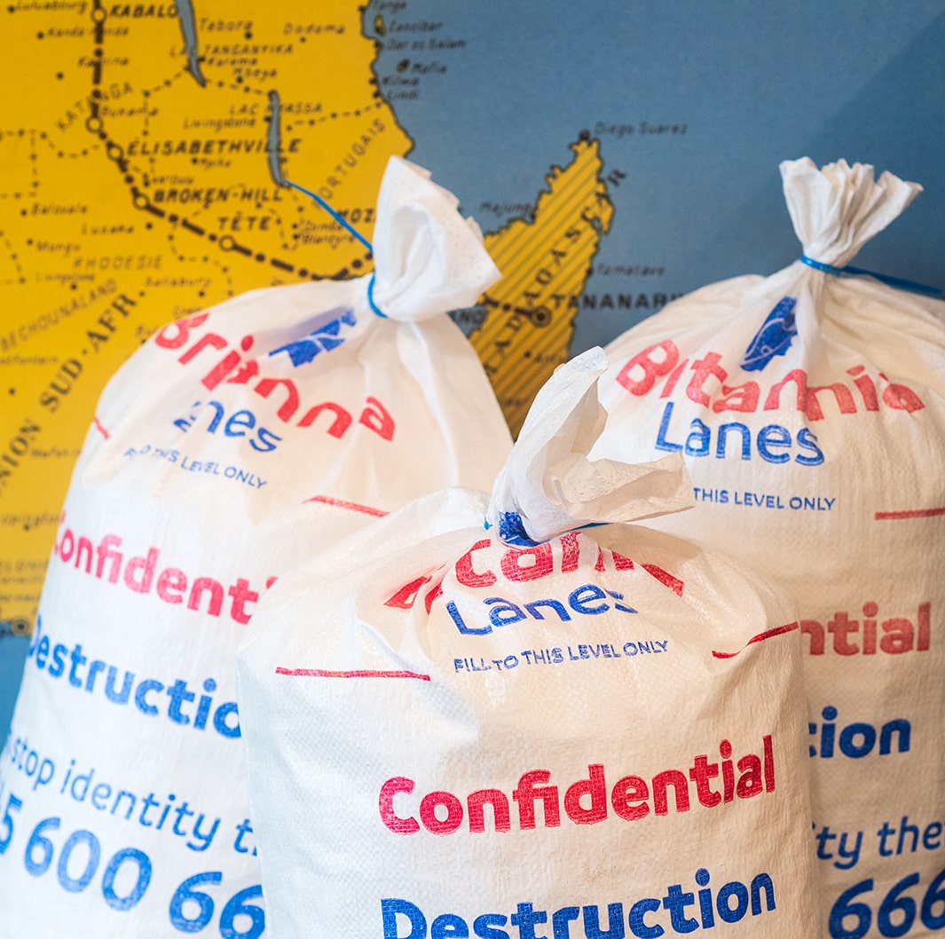 Three white bags labeled "Confidential Destruction" are in the foreground, tied at the top and placed in front of a blue and yellow map. The bags have red and blue text and are from a company named "Britannia Lanes." The map behind them shows part of a coastline.